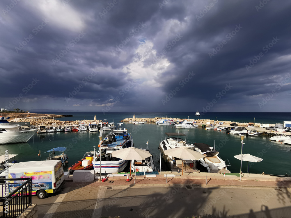 Pier, port with small ships, yachts and boats in the bay of the Mediterranean Sea against the backdrop of a dramatic sky.