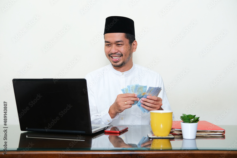 Moslem Asian man smiling while counting his money in front of laptop Moslem Asian man smiling while counting his money in front of laptop