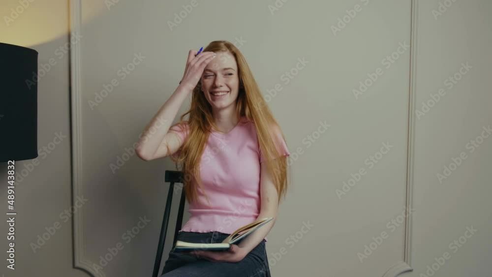 Red-haired female student with copybook smiling at camera doing test task while posing seated on the chair against white wall. Red-haired female student with copybook smiling at camera doing test task while posing seated on the chair against white wall.