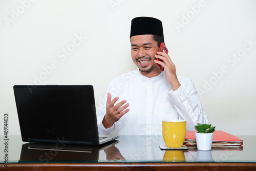 Moslem Asian man smiling when talking in a phone call in front of his laptop photo