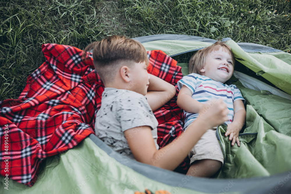 Happy children lying with plaid in camping tent. Family weekend nature outdoor. Smiling teenage boys with mug in hands. Slow life Happy children lying with plaid in camping tent. Family weekend nature outdoor. Smiling teenage boys with mug in hands. Slow life