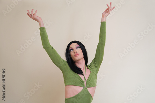 Portrait of a drag queen person looking up with arms raised wearing a green dress posing on beige background.