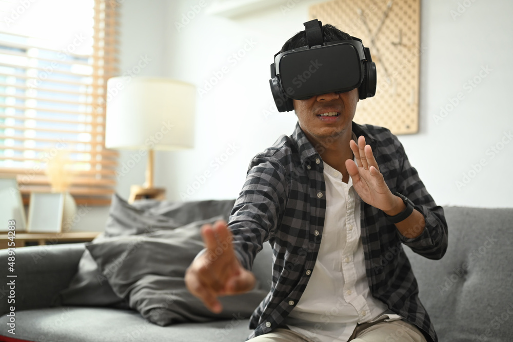 Young asian man sitting on couch and playing fighting video games in VR glass headset.