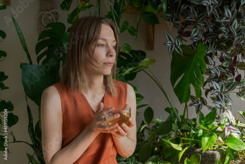 Cute caucasian woman in an orange home dress drinks tea from transparent cup, looking out window against background of indoor green plants, the florist is relaxing at home with enjoying the morning