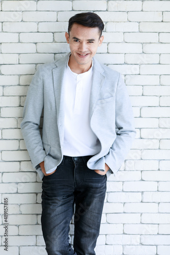 Portrait shot Asian young cool smart handsome muscular undercut hairstyle male businessman in casual gray suit jacket standing holding hands in jeans pockets look at camera on brick wall background