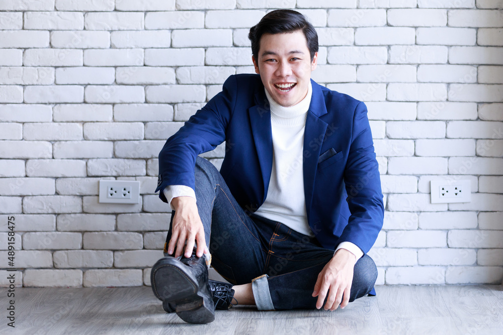 Portrait studio shot Asian fashionable handsome successful professional male businessman in casual blue suit turtle neck shirt sit stretch legs on floor look at camera on white brick wall background