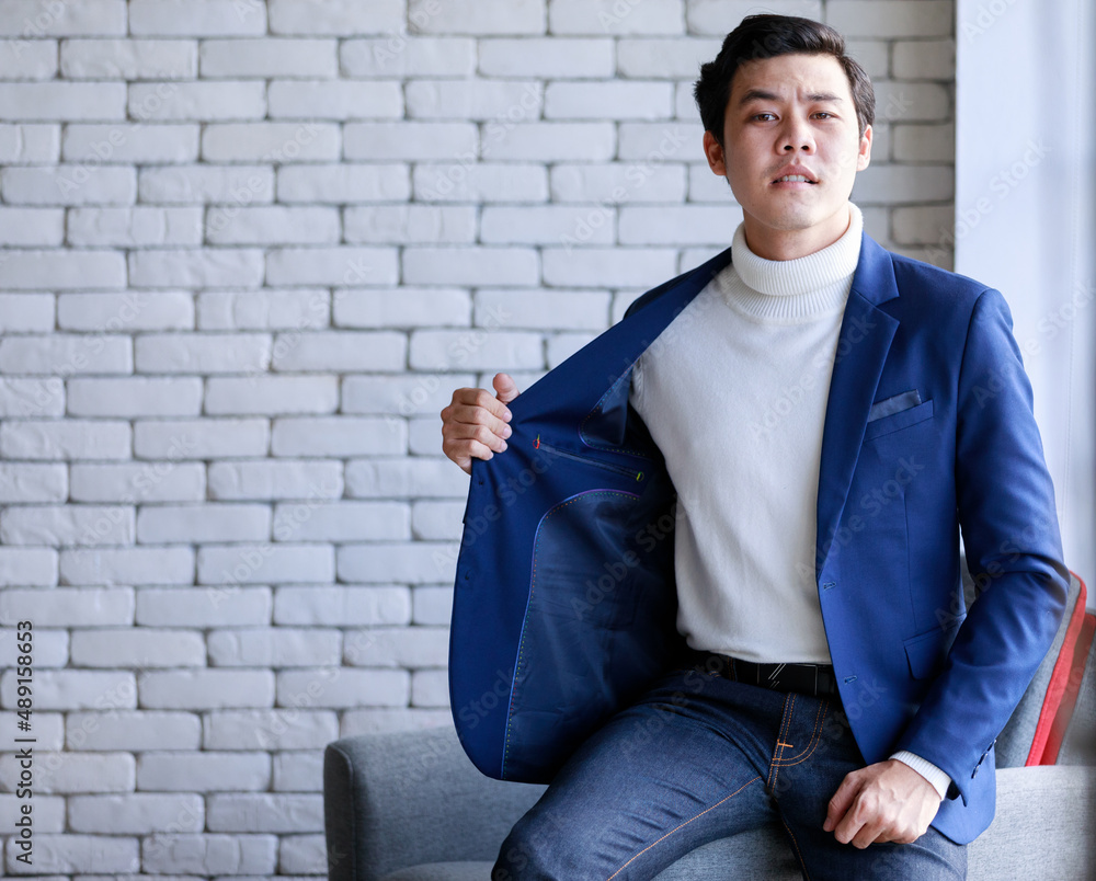 Studio shot Asian young smart handsome successful professional male businessman in casual blue suit turtle neck shirt sit on armchair looking at camera on brick wall background