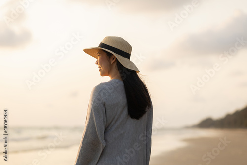 Young asian woman with straw hat standing alone on empty sand beach at sunset seashore. Chilling in holiday weekend summertime. Traveler Female walking around the beach with the sunlight