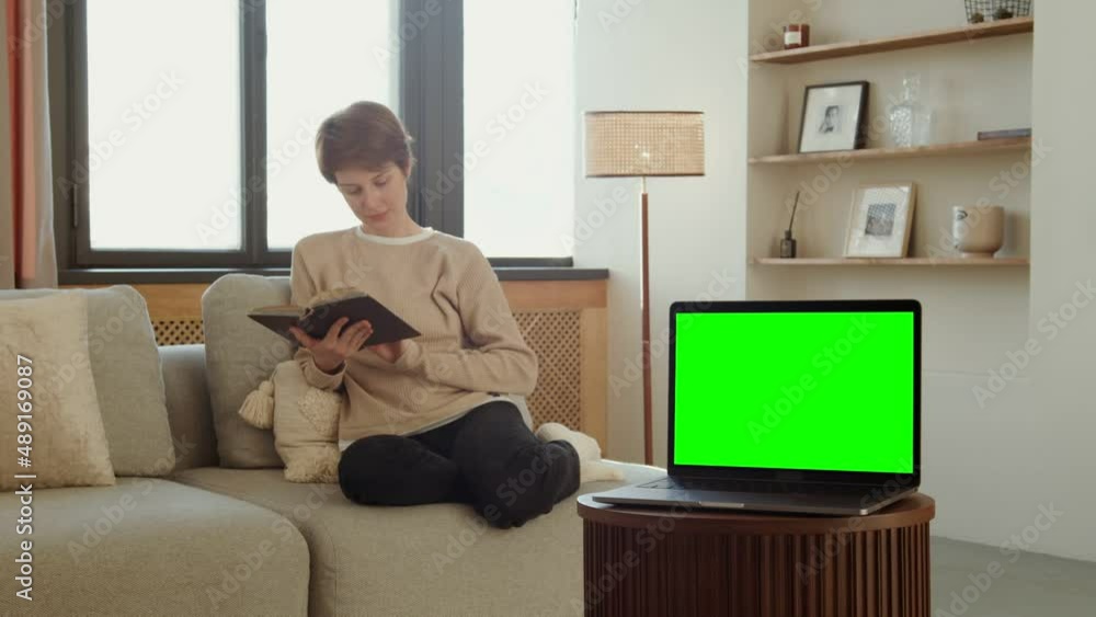 A laptop with a green screen stands on a table against of a woman reading a book A laptop with a green screen stands on a table against of a woman reading a book
