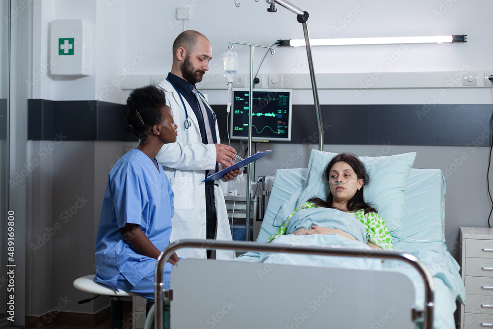 Obraz premium Recovering woman on hospital bed with iv drip line and vital functios monitored being consulted by doctor with medical history on clipboard. medic doing consult with nurse looking at patient chart.