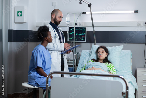 Recovering woman on hospital bed with iv drip line and vital functios monitored being consulted by doctor with medical history on clipboard. medic doing consult with nurse looking at patient chart.