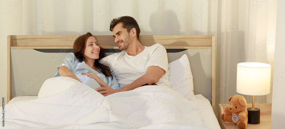 Fototapeta premium Happy Caucasian adult couple laugh and smile while talking fun and embrace each together on bed as enjoy resting under warm quilt to sleep in bedroom on romantic night at home