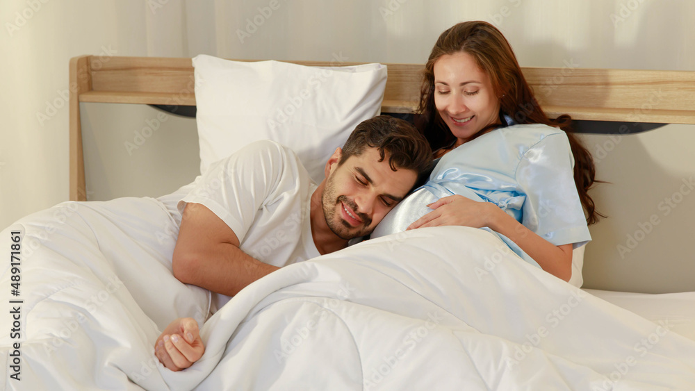 Fototapeta premium Delighted father tenderly lie down on belly of beloved pregnant wife on bed to feel and hear unborn baby kicking in stomach
