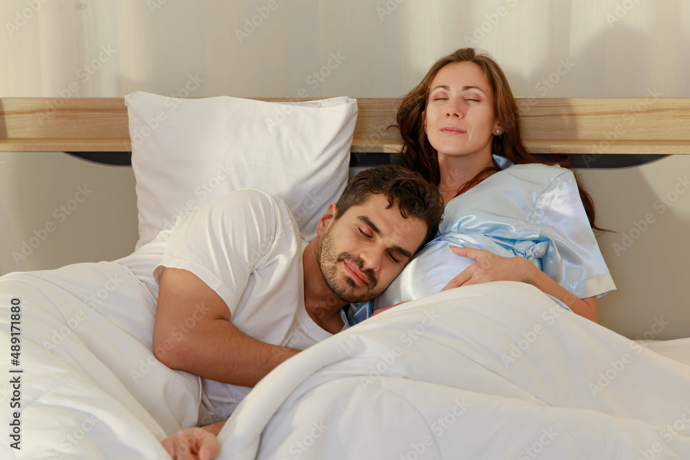 Fototapeta premium Delighted father tenderly lie down on belly of beloved pregnant wife on bed to feel and hear unborn baby kicking in stomach