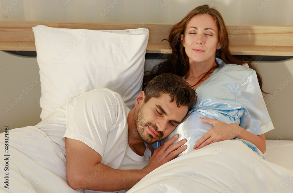 Delighted father tenderly lie down on belly of beloved pregnant wife on bed to feel and hear unborn baby kicking in stomach Delighted father tenderly lie down on belly of beloved pregnant wife on bed to feel and hear unborn baby kicking in stomach
