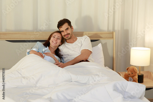 Happy Caucasian adult couple laugh and smile while talking fun and embrace each together on bed as enjoy resting under warm quilt to sleep in bedroom on romantic night at home