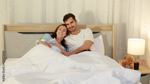 Happy Caucasian adult couple laugh and smile while talking fun and embrace each together on bed as enjoy resting under warm quilt to sleep in bedroom on romantic night at home