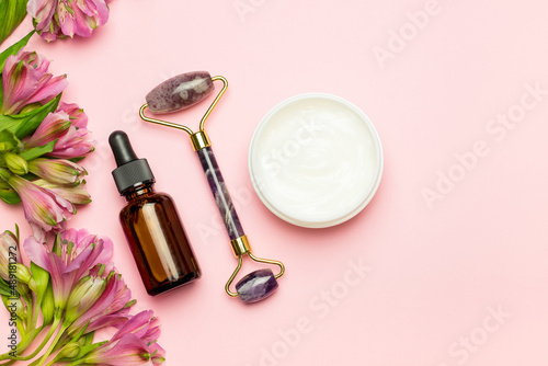 Face roller with cosmetic serum and face cream on a pink background with flowers. Facial massage kit for lifting massage.