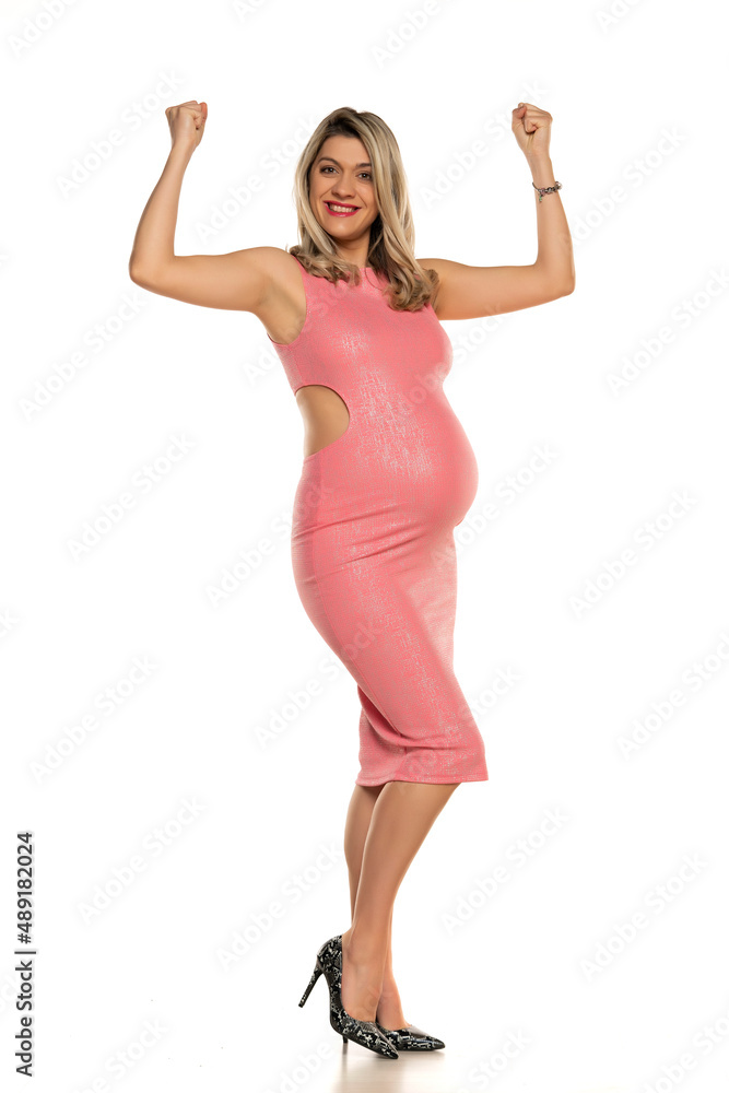 Pregnant woman in tight pink dress showing biceps in studio Pregnant woman in tight pink dress showing biceps in studio