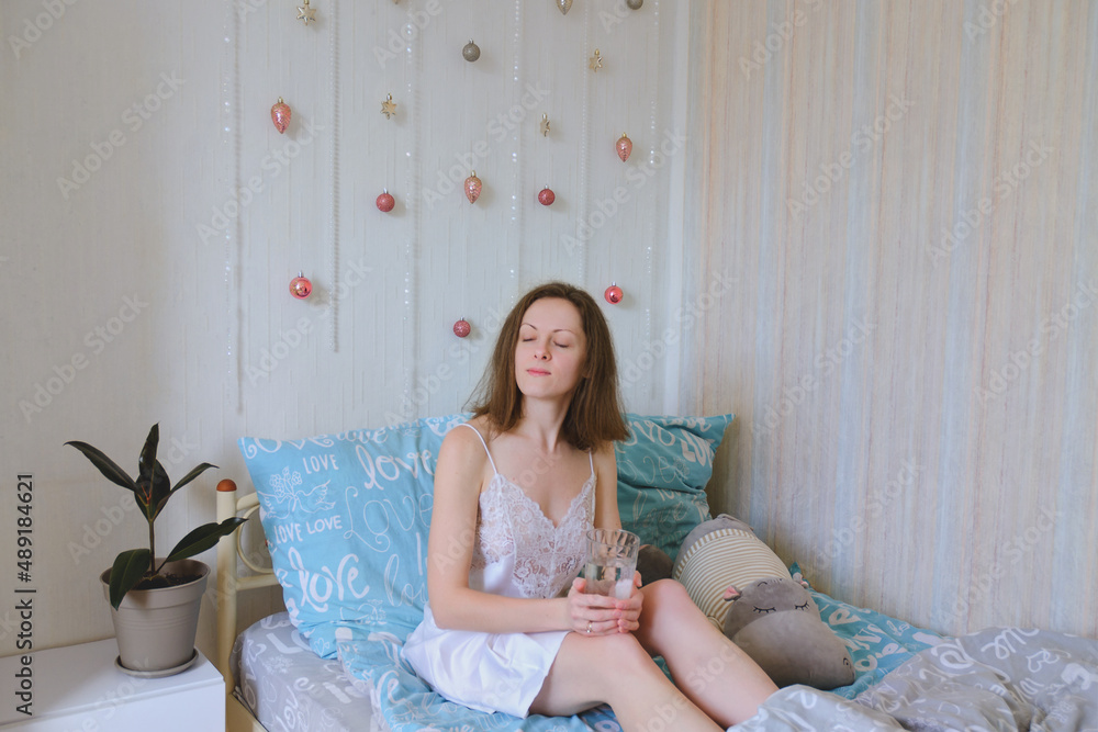 Young happy smiling woman, bride lies in bed in pajamas, bathrobe in a cozy home. Drinking water. Relax. Healthy lifestyle. Morning rituals. Young happy smiling woman, bride lies in bed in pajamas, bathrobe in a cozy home. Drinking water. Relax. Healthy lifestyle. Morning rituals.