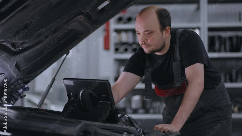 Male Mechanic Uses a Tablet Computer with an Augmented Reality Diagnostics Software. Specialist Inspecting the Car in Order to Find Broken Components Inside the Engine Bay. Modern Car Service. Male Mechanic Uses a Tablet Computer with an Augmented Reality Diagnostics Software. Specialist Inspecting the Car in Order to Find Broken Components Inside the Engine Bay. Modern Car Service.