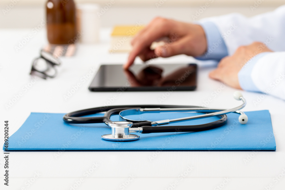Closeup shot of a stethoscope resting on a tablet with an unrecognizable doctor working in the background with laptop Closeup shot of a stethoscope resting on a tablet with an unrecognizable doctor working in the background with laptop