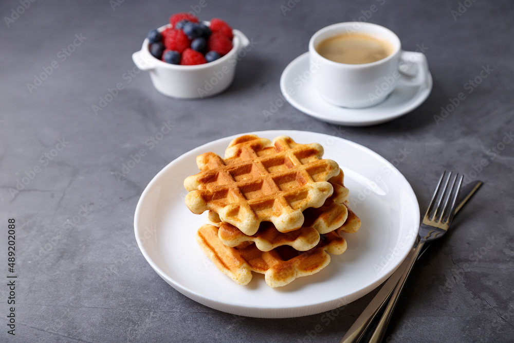 Viennese or Belgian waffles with fresh berries (raspberries and blueberries) on a white plate and a cup of coffee. Traditional dessert. Close-up, grey background. Viennese or Belgian waffles with fresh berries (raspberries and blueberries) on a white plate and a cup of coffee. Traditional dessert. Close-up, grey background.