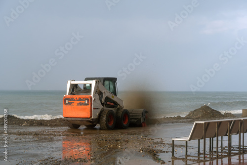 Small tractor with a large brush cleans the pedestrian, coastal area from dirt and sand after a storm against the background of a raging sea. Batumi, Georgia