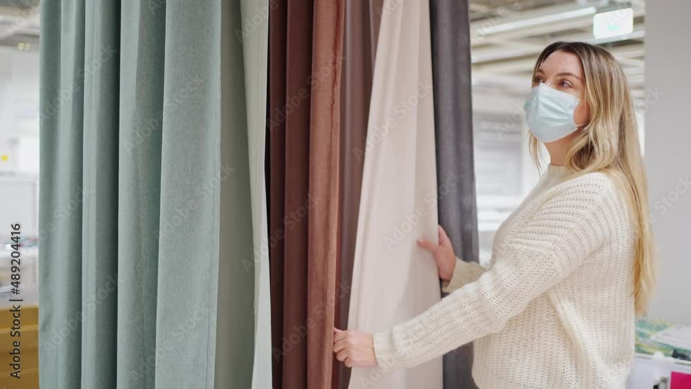 young woman in a protective mask chooses fabric and curtains in a home design store young woman in a protective mask chooses fabric and curtains in a home design store