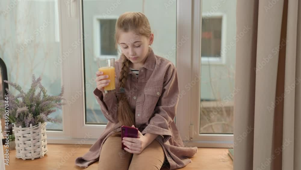 girl drinking juice with a phone in her hands sitting on the windowsill