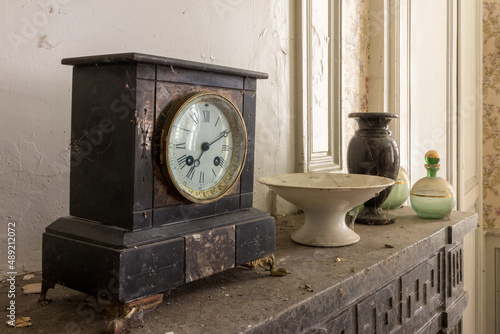 Old clock on a chimney with a lot of dust and dirt in an abandoned house. In the background are old photo