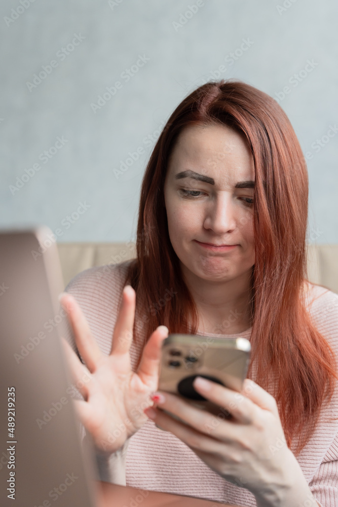 Confused young woman with red hair sitting on sofa and talkink to someone by video call. Confused young woman with red hair sitting on sofa and talkink to someone by video call.