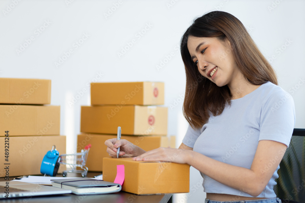 Young Asian woman running a small e-commerce start-up is working at her office desk and is writing the shipping address on a parcel for a customer. Online shopping concept. Young Asian woman running a small e-commerce start-up is working at her office desk and is writing the shipping address on a parcel for a customer. Online shopping concept.