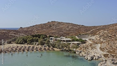Paros Greece Aerial v1 dolly in shot toward sandy monastiri beach with umbrellas on the shore, nature reserve park on rocky coast and rocky hill - September 2021 photo
