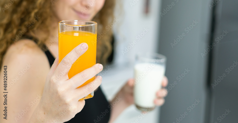 orange juice glass in hand of healthy woman while holding milk glass in other hand in kitchen with space. beautiful caucasian woman cooking healthy diet fruit or vegetable in kitchen orange juice glass in hand of healthy woman while holding milk glass in other hand in kitchen with space. beautiful caucasian woman cooking healthy diet fruit or vegetable in kitchen