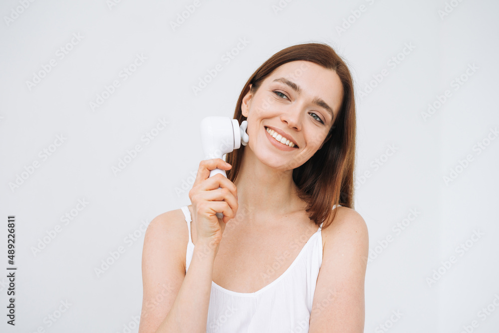 Beauty portrait of happy smiling woman with dark long hair with facial massager in hand on clean fresh skin face on the white background isolated Beauty portrait of happy smiling woman with dark long hair with facial massager in hand on clean fresh skin face on the white background isolated