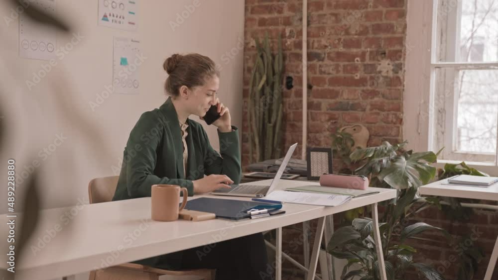 Medium slowmo of young Caucasian businesswoman talking on smartphone sitting in front of laptop at her workplace in modern office Medium slowmo of young Caucasian businesswoman talking on smartphone sitting in front of laptop at her workplace in modern office