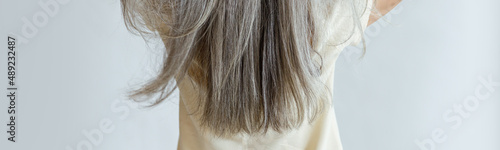 Middle aged woman in yellow blouse shows long loose grey hair standing on light background in studio backside view. Mature beauty lifestyle