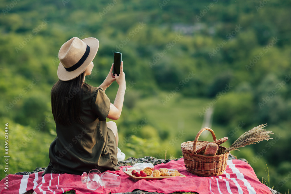 Young girl with hat making photo on smartphone at beautiful landscape while resting at summer picnic