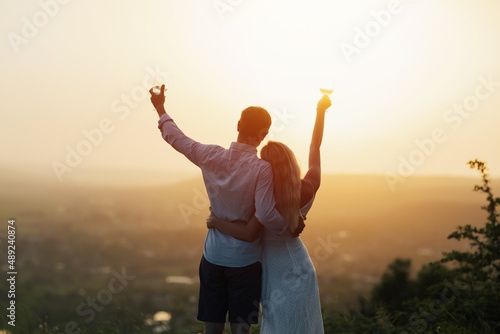 Beautiful young caucasian couple drinking wine while standing back at camera during date on hill in evening. Romantic date concept