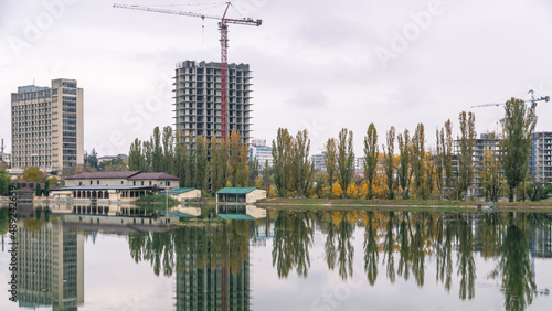 High-rise buildings are being built next to the pond. Reflection of buildings and trees under construction in the lake water. Construction of high-rise buildings. Tower cranes near new buildings.
