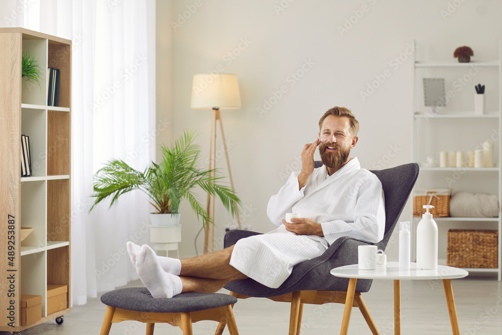 Portrait of happy handsome unshaven ginger man in white bathrobe and fresh clean socks having beauty day at home, enjoying skin care, relaxing in comfy chair, applying face cream, drinking detox tea Portrait of happy handsome unshaven ginger man in white bathrobe and fresh clean socks having beauty day at home, enjoying skin care, relaxing in comfy chair, applying face cream, drinking detox tea