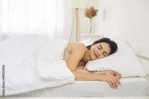 Happy tranquil beautiful woman napping lying on comfortable bedding in bedroom at home alone. Side view of smiling caucasian woman enjoying resting. Refreshment, renew energy and strength concept.