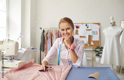 Portrait of smiling young dressmaker or tailor sew fabric create new garment in own workshop. Happy designer or seamstress cut design clothing for client in fashion atelier. Style concept.
