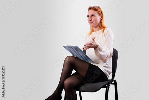 A girl, in a white blouse, sits on a chair with a folder in her hands, on a gray background
