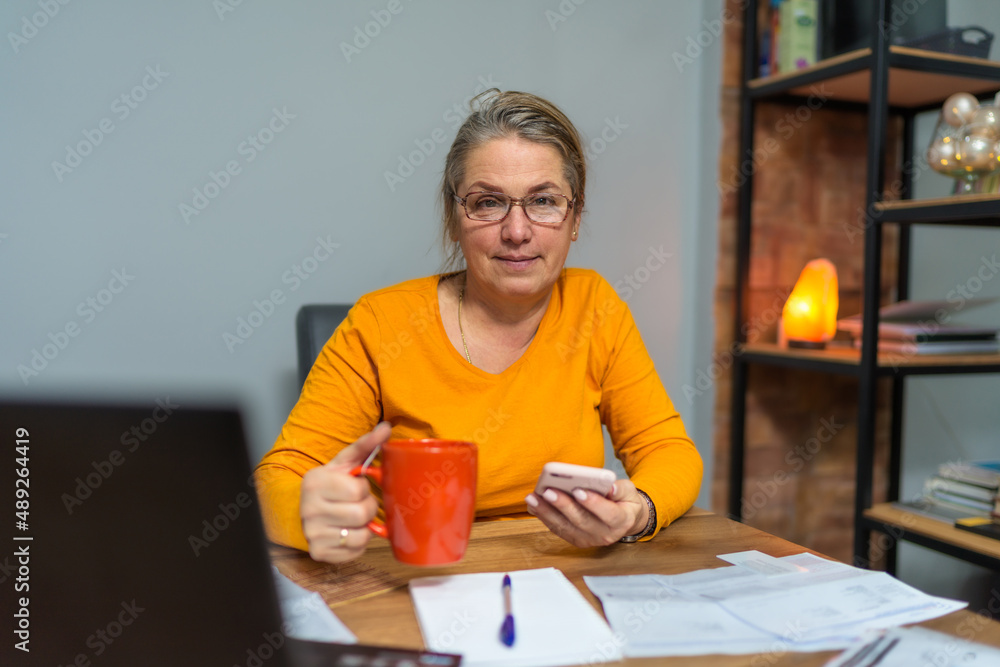 An older mature business woman who keeps a paper account using a mobile phone and laptop, an old woman manages the finances of the account, calculates the budget tax. An older mature business woman who keeps a paper account using a mobile phone and laptop, an old woman manages the finances of the account, calculates the budget tax.