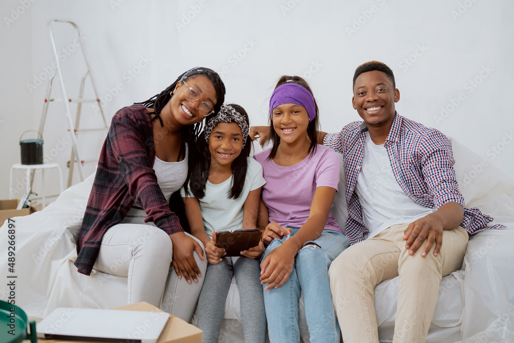Portrait of a happy smiling loving family sitting on a couch during an apartment renovation. Young parents and two daughters browsing room inspirations on a tablet Portrait of a happy smiling loving family sitting on a couch during an apartment renovation. Young parents and two daughters browsing room inspirations on a tablet