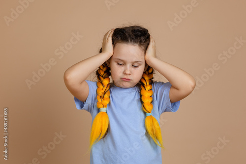 Upset, Caucasian small female child holding head with hands looking down having kanekalon braids of yellow color on head in blue t-shirt on beige background.