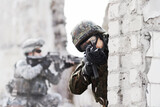 Dynamic defense force. A soldier peering around a wall while pointing a gun at the camera during a rade on a damaged building.