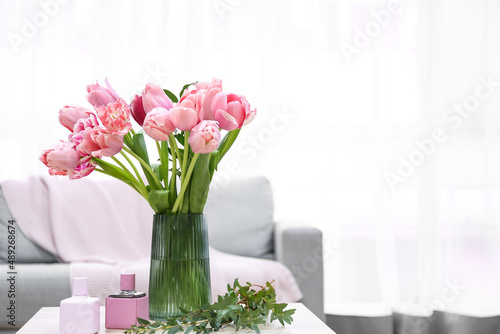 Vase with pink tulips, bottles of perfume and plant branches on table in living room